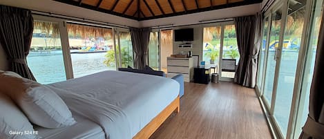 Un dormitorio moderno con una cama grande, un escritorio y un televisor. Tiene ventanas amplias con vistas a un cuerpo de agua y un parque infantil.