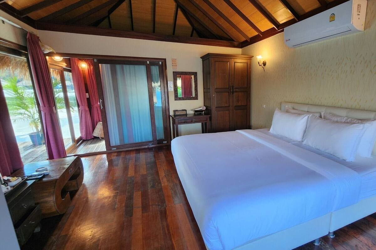 A bedroom with a large bed, wooden flooring, a desk with a chair, a wardrobe, and an air conditioning unit.