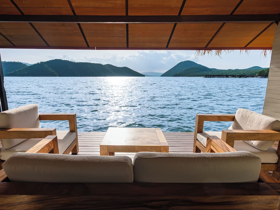 Una terraza de madera con sofá y sillones frente a un cuerpo de agua y colinas al fondo.
