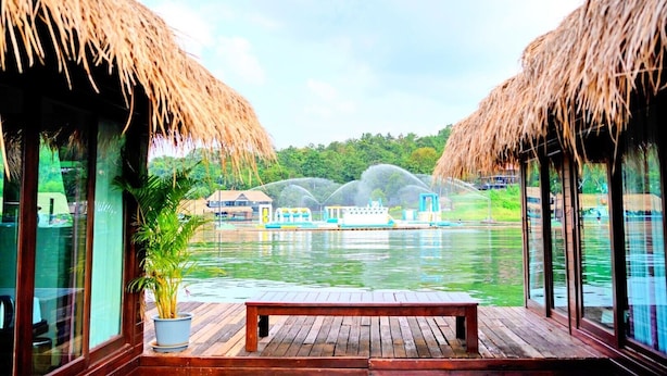 A wooden deck with a bench, a potted plant, and a view of a water body with boats and a building in the background. - Ruknam Resort (Si Sawat)