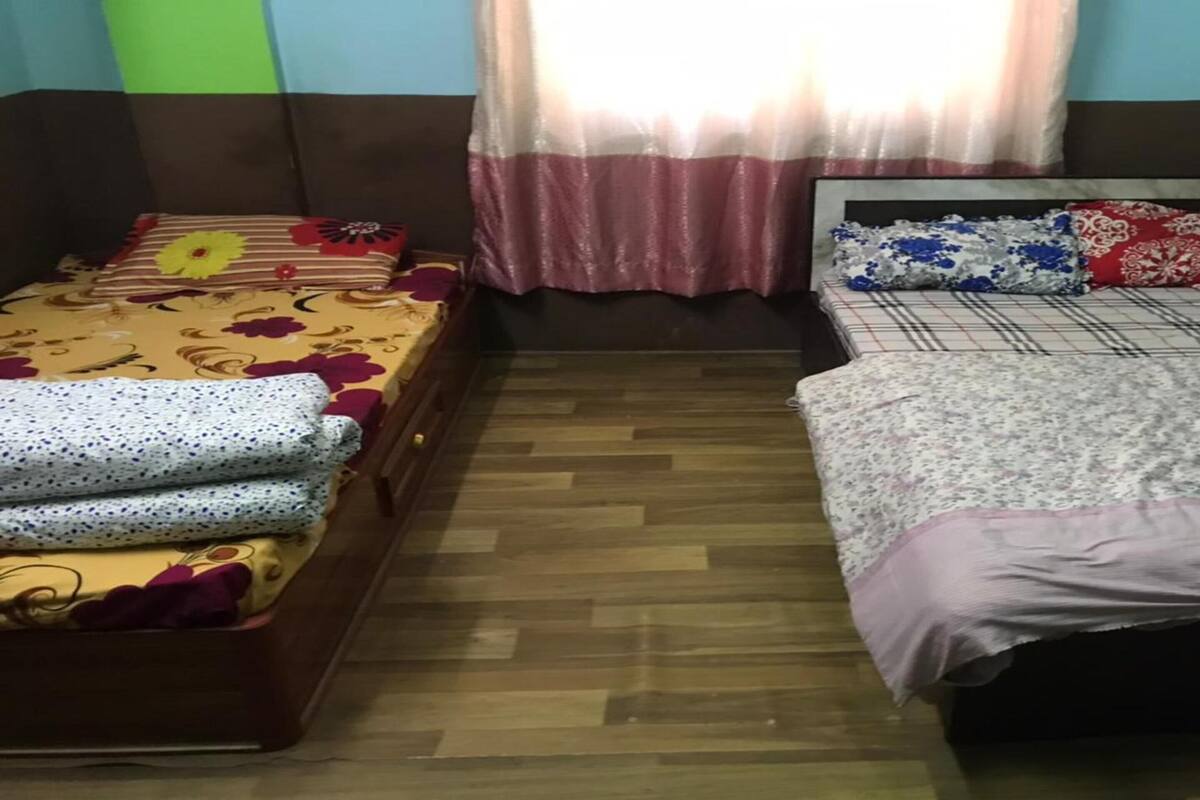 Ein Zimmer mit zwei Betten, eines mit einer Blumenbettdecke und das andere mit einer karierten Bettdecke. Es gibt ein Fenster mit transparenten Vorhängen, und der Boden ist aus Holz.