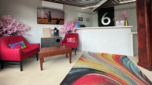 Una hall d'albergo con una reception, una poltrona rossa, un tavolino in legno, un tappeto colorato e una ventola a soffitto.