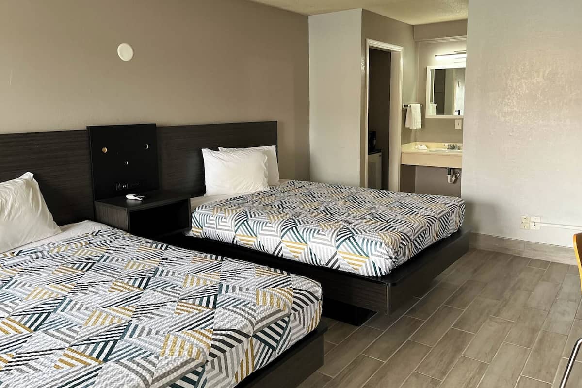 Hotellihuone, jossa on kaksi sänkyä, tumma puinen pääty, yöpöytä, kylpyhuone peilillä ja pesualtaalla sekä laattalattia.