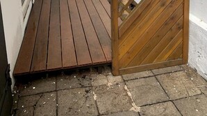 Un espacio exterior estrecho con una terraza de madera, un muro de ladrillo blanco y un portón de madera.