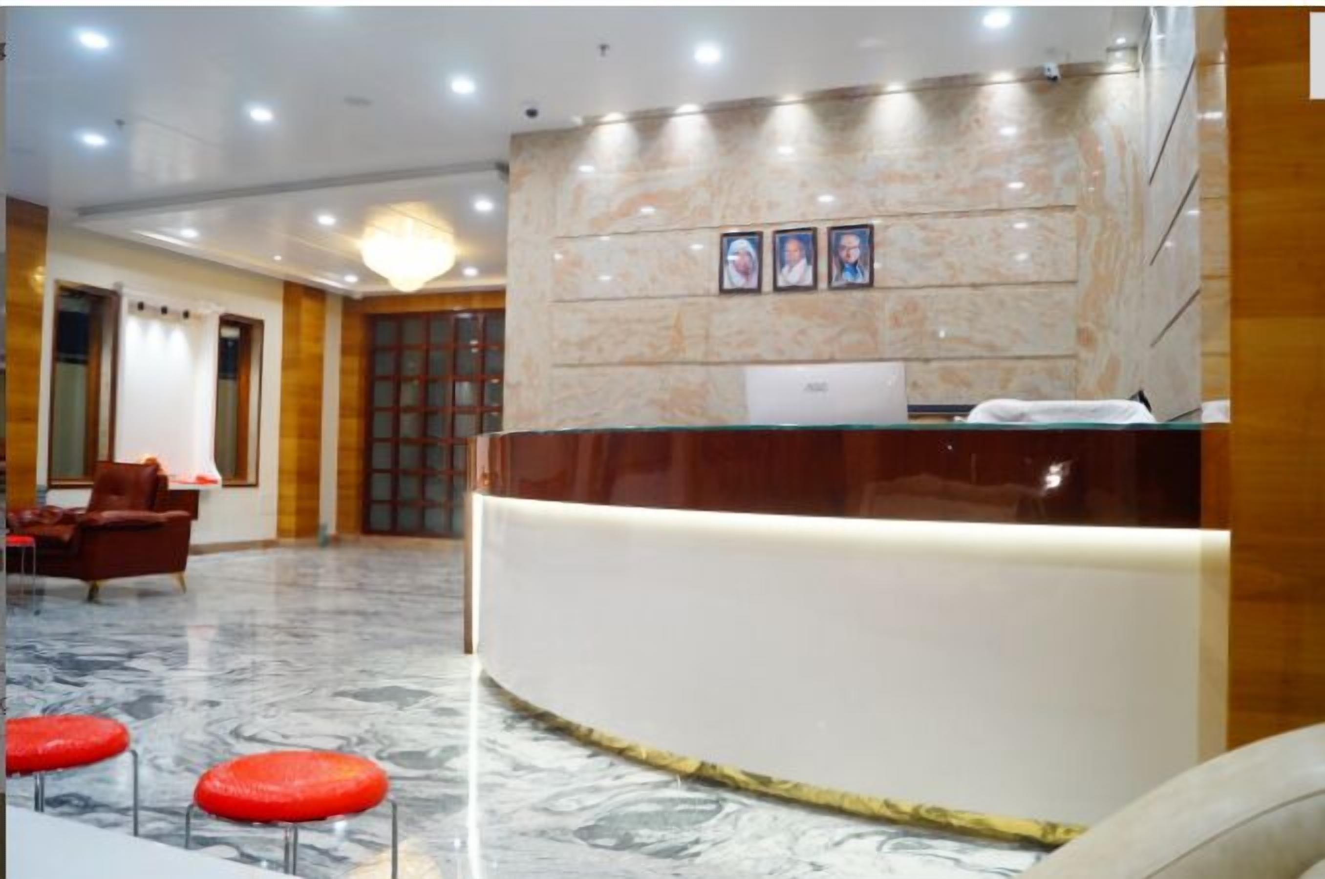 A hotel reception area with a marble floor, a curved reception desk, and a seating area with red stools.