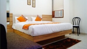 Een net opgemaakt bed met witte lakens en oranje kussens, een houten hoofdbord, een klein nachtkastje en een stoel.