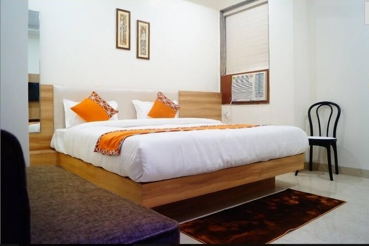 Een net opgemaakt bed met witte lakens en oranje kussens, een houten hoofdbord, een klein nachtkastje en een stoel.
