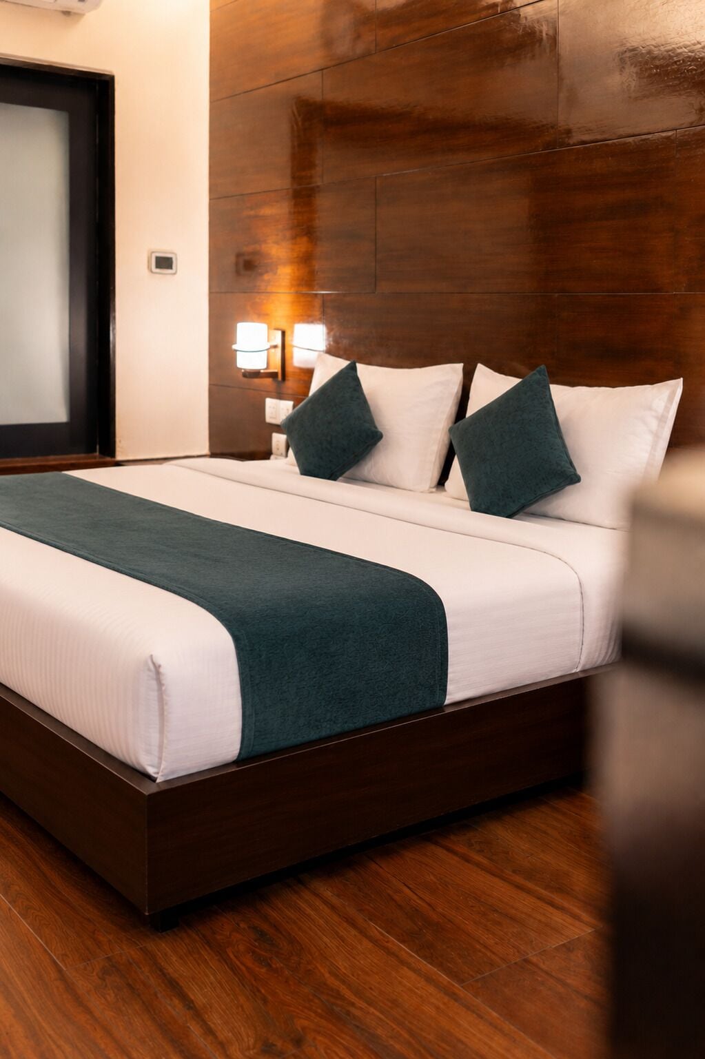 A hotel room with a wooden headboard, a bed with white and teal bedding, and a nightstand with a lamp.