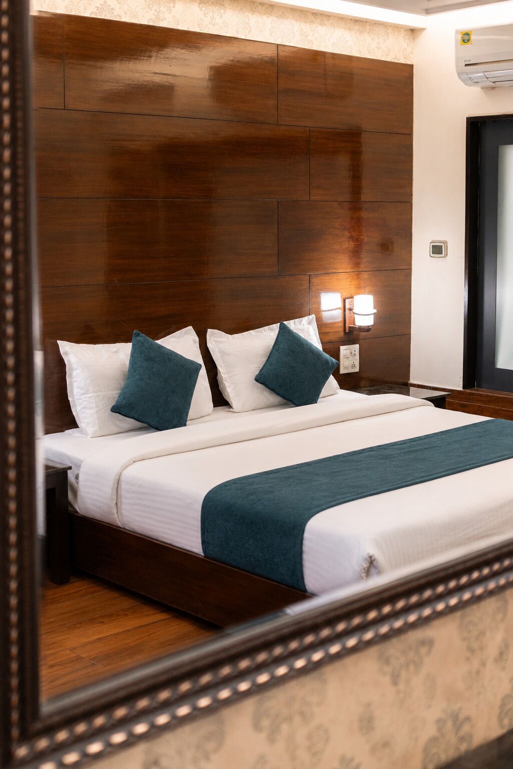 A hotel room with a wooden headboard, a bed with white bedding and teal pillows, a bedside table, and a wall-mounted light fixture.