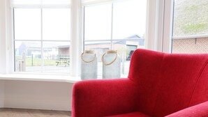 Ein roter Sessel in einem Raum mit großen Fenstern, Blick auf ein Gebäude im Freien und zwei Korbwannen am Fensterbrett.