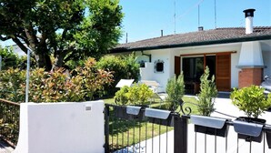Une maison à deux étages, à l’extérieur blanc, avec une cheminée en briques et un petit jardin devant, composé d’une pelouse et de quelques plantes en pot.