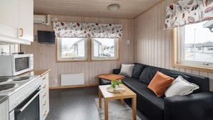 Un espacio habitable compacto con kitchenette, un sofá con cojines, una mesita con una planta y una ventana con vistas a una zona residencial.