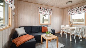 Interior acogedor de una cabaña con un sofá negro, una mesa de comedor de madera, sillas blancas y una alfombra pequeña.