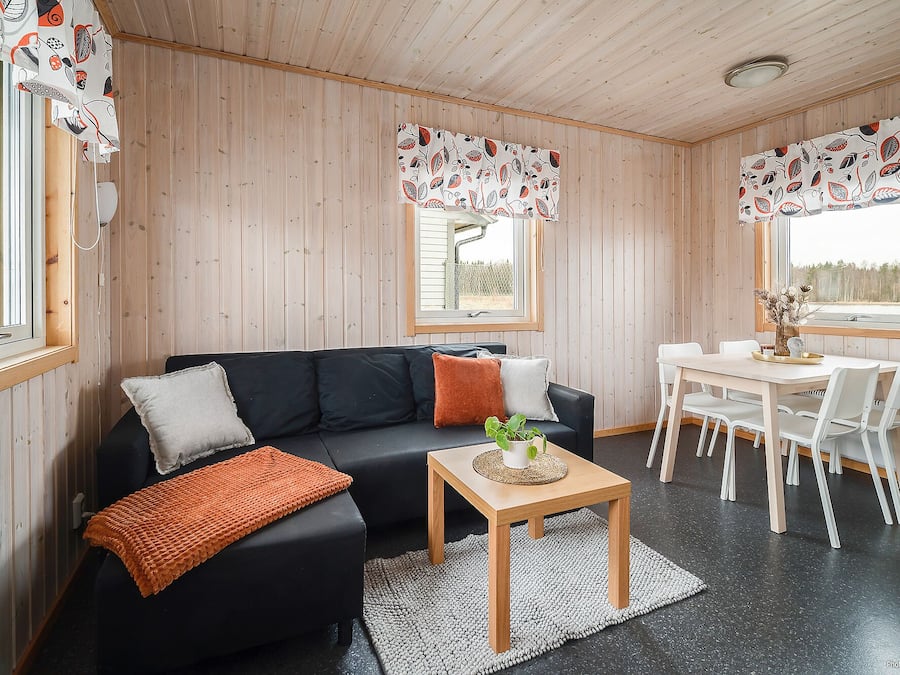 En mysig stuga med en svart soffa, ett matbord i trä, vita stolar och en liten matta.
