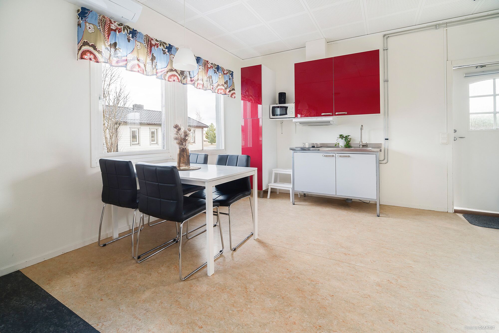 Un comedor con mesa y sillas, una cocina con armarios rojos y una ventana con vista a casas.