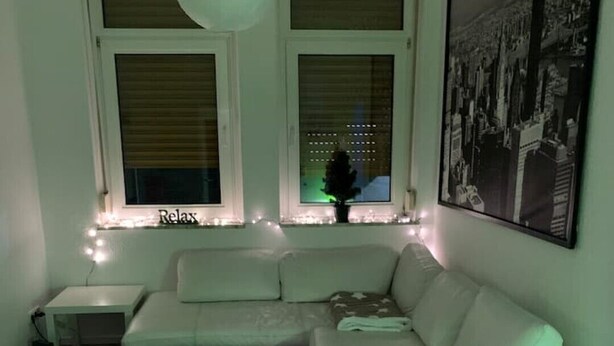 A modern living room with a white sofa, a coffee table, and a side table. There are two windows with blinds, a hanging light fixture, and a framed picture on the wall.