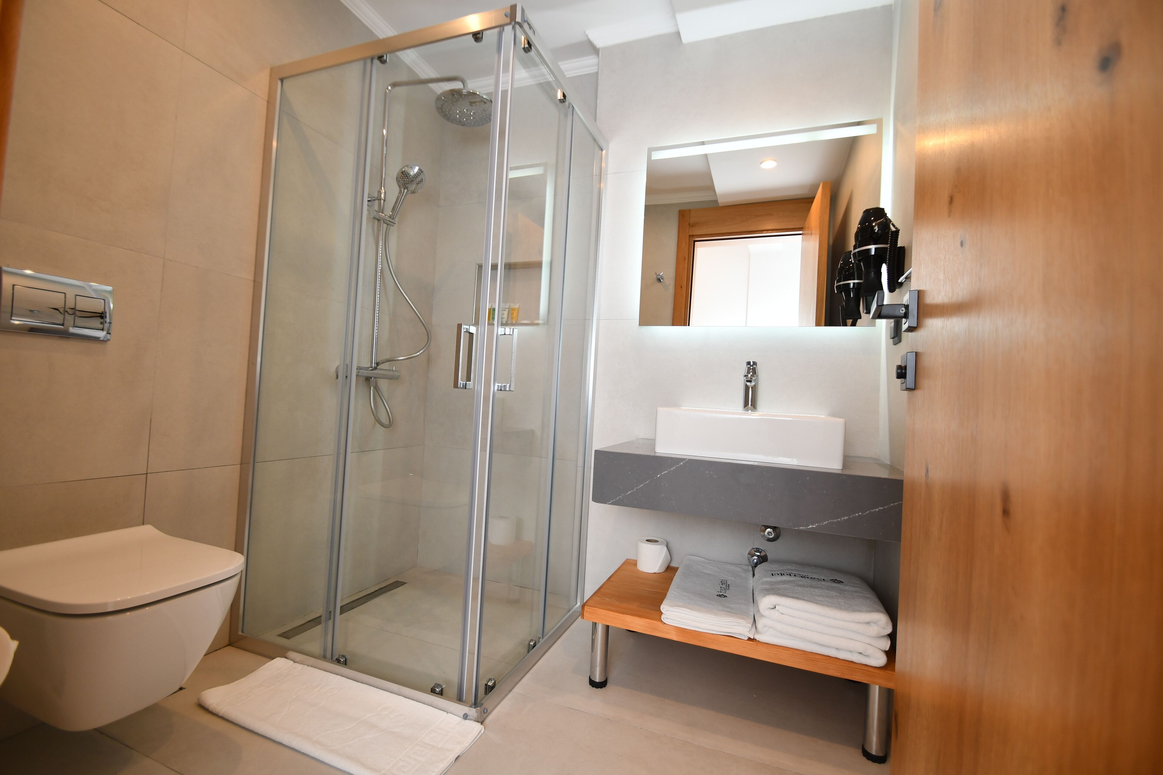 A modern bathroom with a glass-enclosed shower, a toilet, a sink with a mirror, and a wooden shelf with towels.