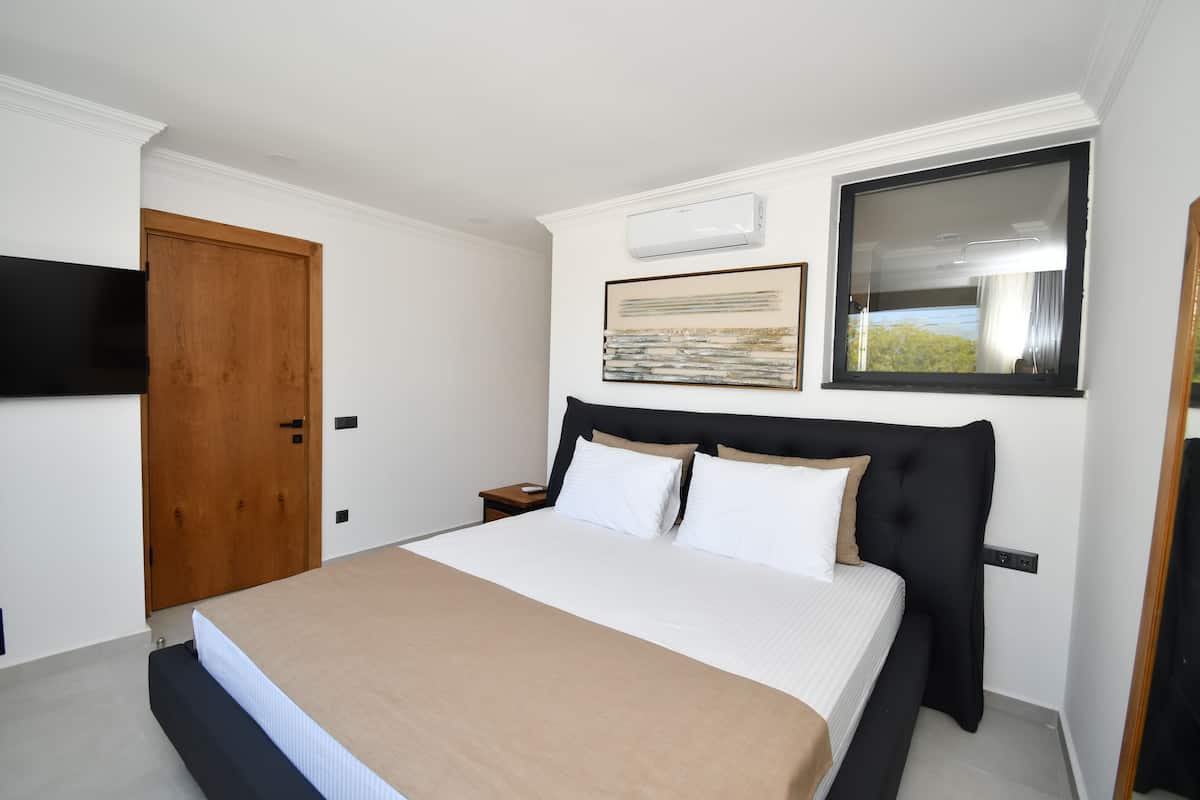 A bedroom with a bed, a wooden door, a nightstand, a wall-mounted TV, and a window with a view of greenery.