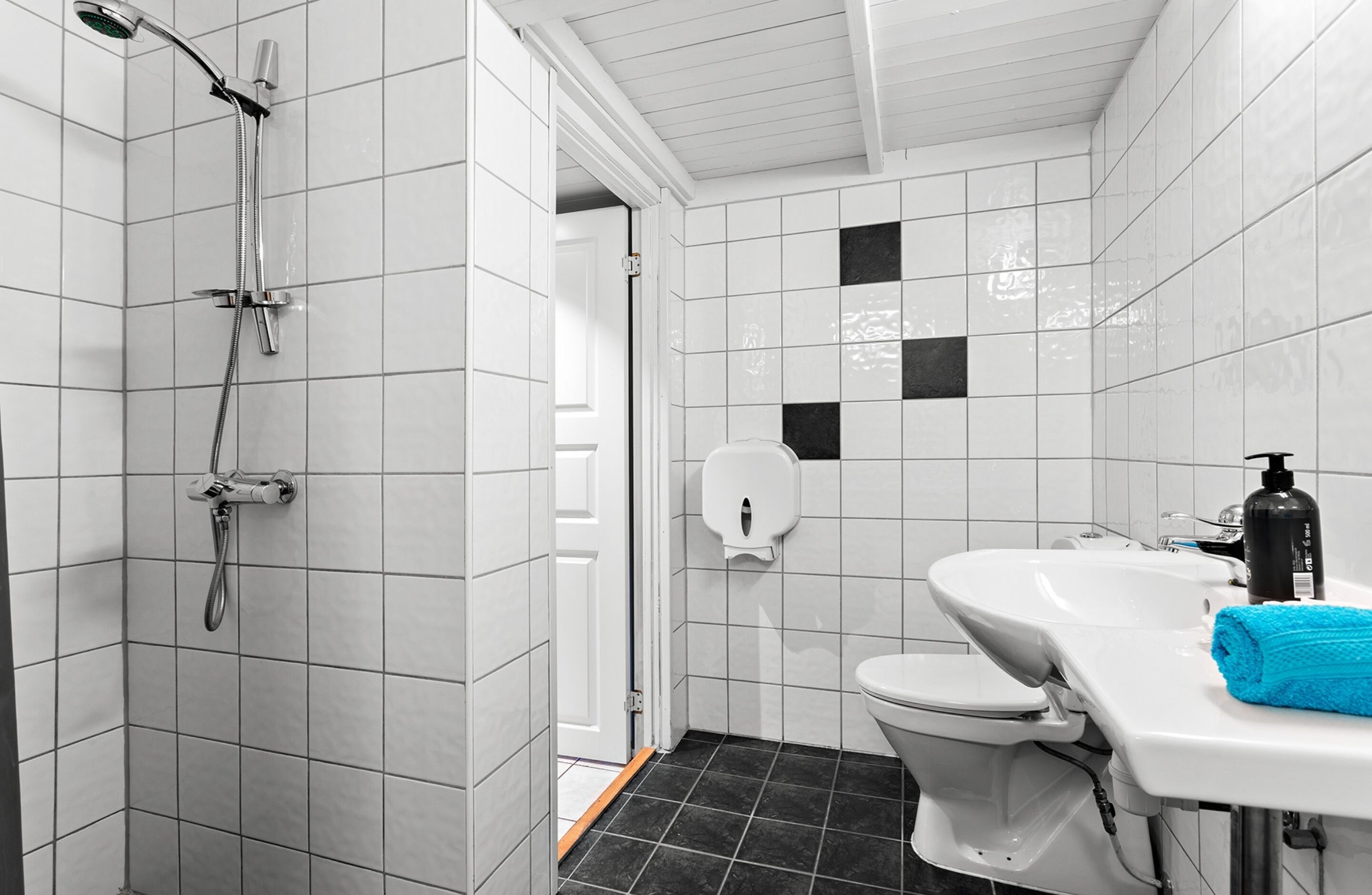 A bathroom with white tiled walls, a black tiled floor, a shower, a toilet, and a sink with a blue towel.