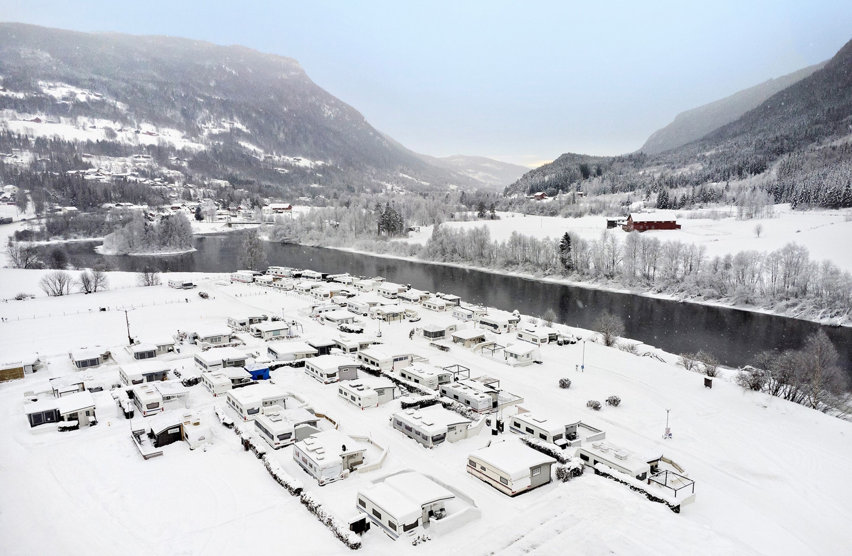 A snow-covered RV park with multiple RVs parked in an open area, surrounded by snow-covered trees and mountains.