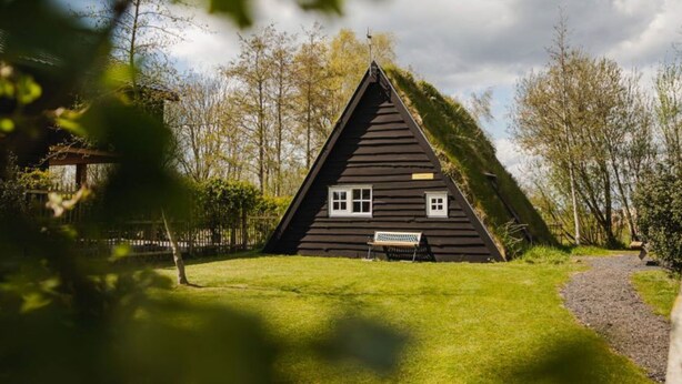 Ein dreieckiges Holzhaus mit einer Bank davor, umgeben von Bäumen und einem wolkenlosen Himmel.