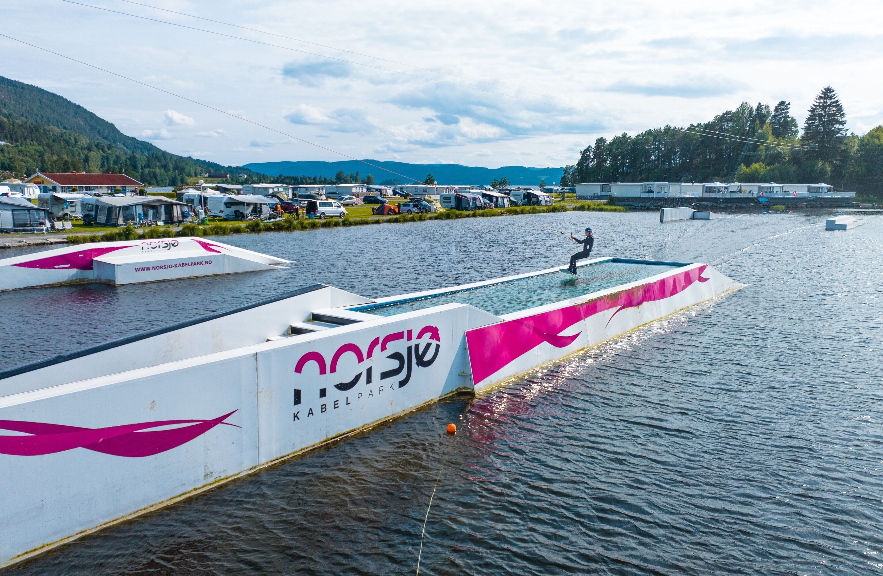 A water-based recreational area with a white structure featuring the text "Norsjo KABELPARK" and a person standing on a platform.