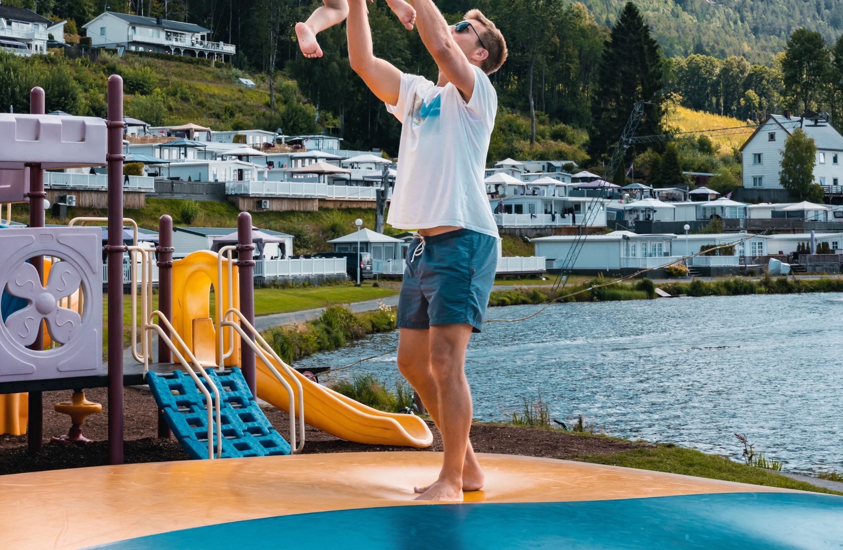 Una persona haciendo un pino en un parque infantil junto a un lago, con casas al fondo.