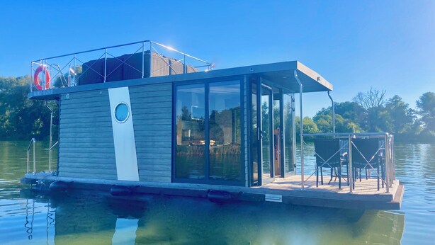 Una moderna casa flotante con cerramiento de cristal, terraza con asientos y un flotador en el techo.