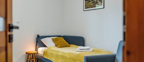 A small, neatly arranged bedroom with a single bed, a chair, a small stool, and a framed picture on the wall.