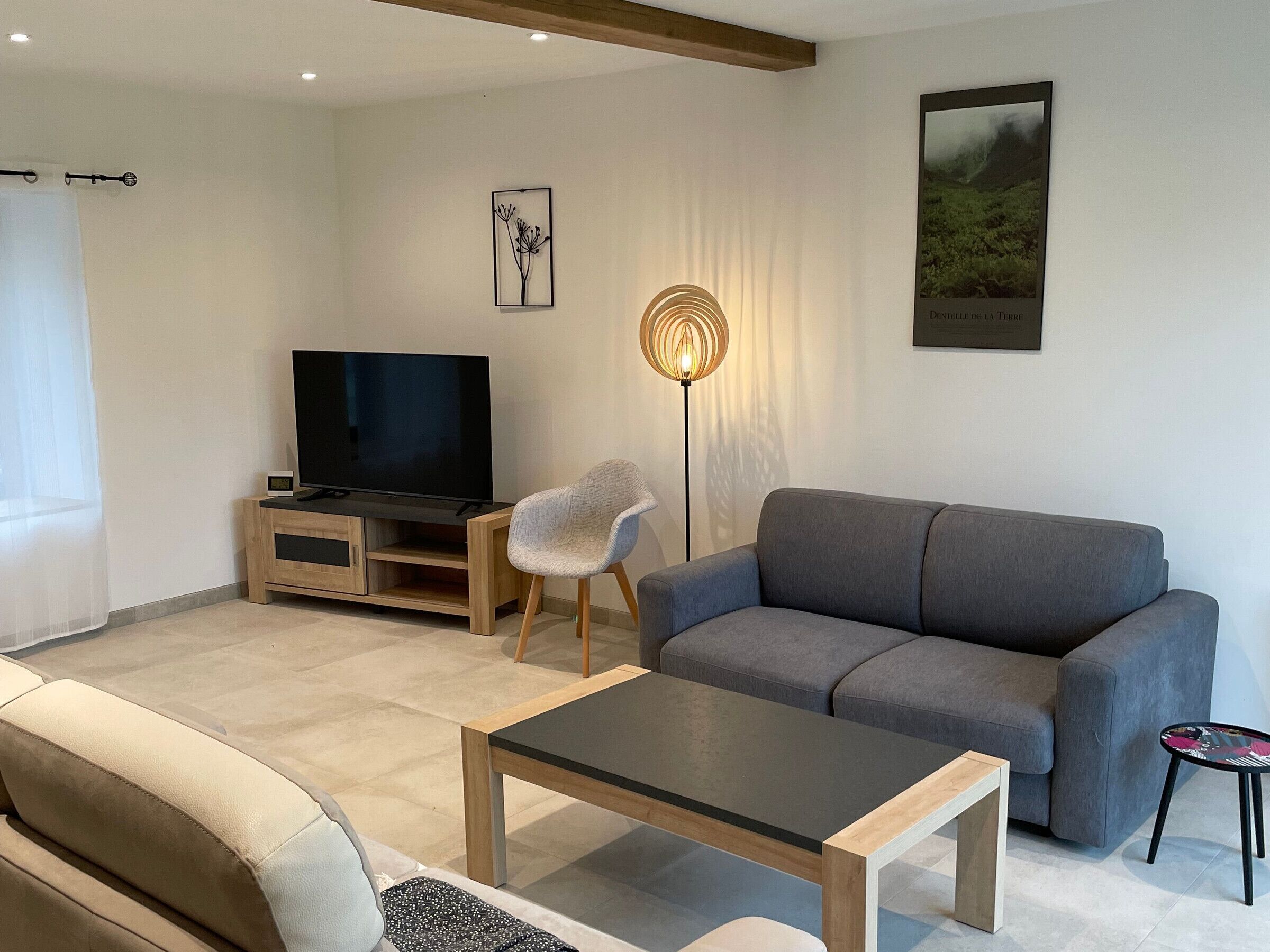 Ett modernt vardagsrum med en grå soffa, ett soffbord i trä, en tv på ett träställ, en golvlampa och en inramad tavla på väggen.