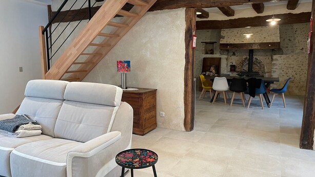 Una sala moderna con un sofá, un taburete, una escalera de madera y un comedor con sillas y una mesa.