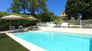Piscina con sillas de descanso y sombrillas, rodeada por una cerca y follaje.