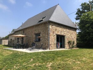 Um edifício de pedra com telhado inclinado, um pátio com mobiliário para área externa e um pequeno jardim.