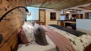 Un dormitorio rústico con techo de madera, cama con ropa de cama a cuadros, una lámpara de noche y vistas hacia una zona de cocina.