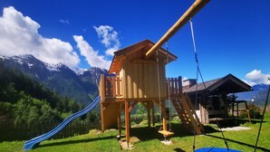 Una casita de juegos de madera con un tobogán y un columpio en un área cubierta de césped, con montañas al fondo.