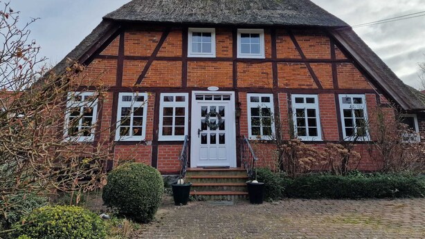 Une maison traditionnelle à colombages, dotée d’un toit de chaume à forte pente, d’une porte blanche et de nombreuses fenêtres.