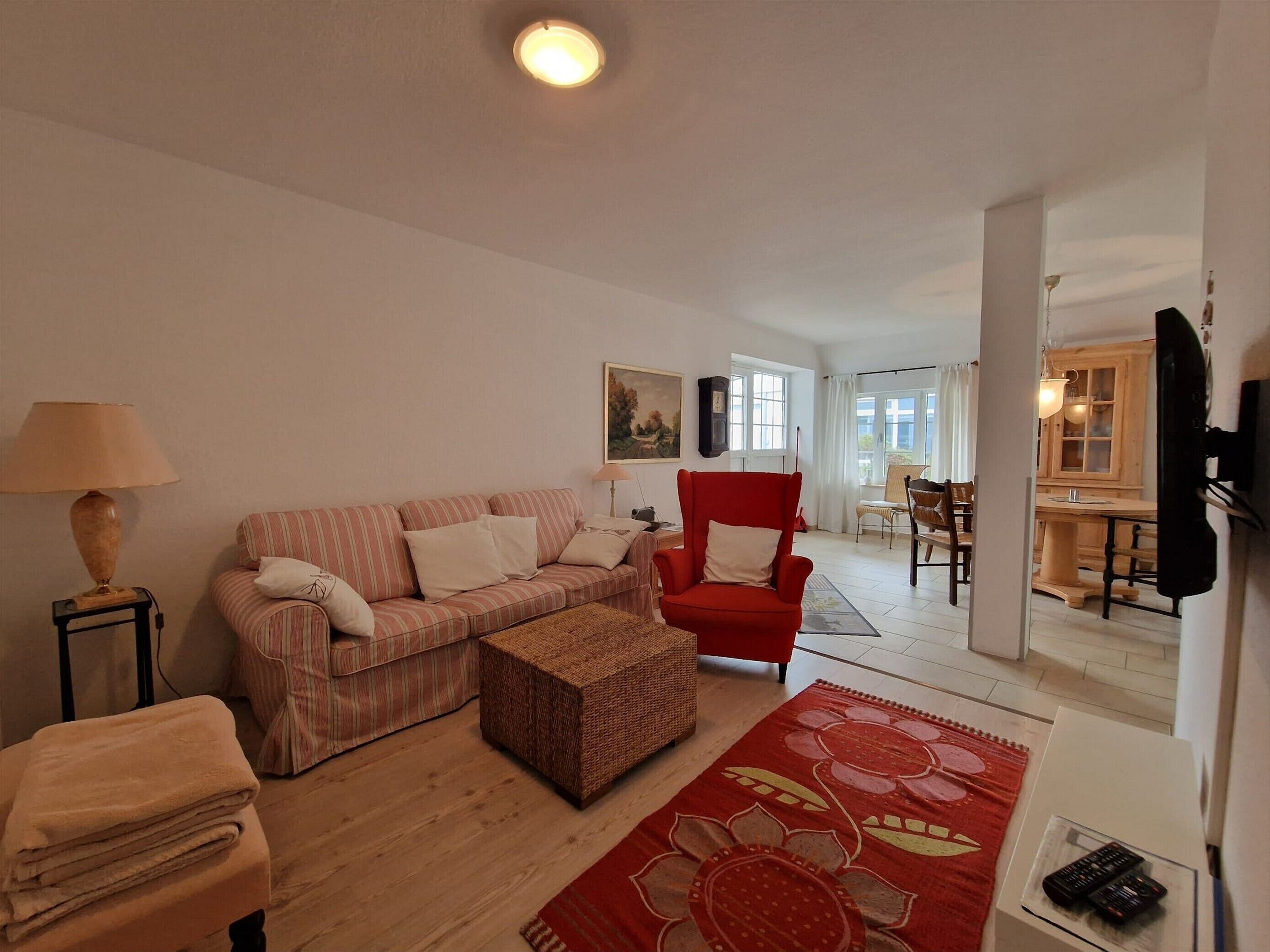 Uma sala de estar com um sofá listrado, uma poltrona vermelha, um banquinho trançado, um tapete floral vermelho e uma luminária. É possível ver uma área de jantar ao fundo.