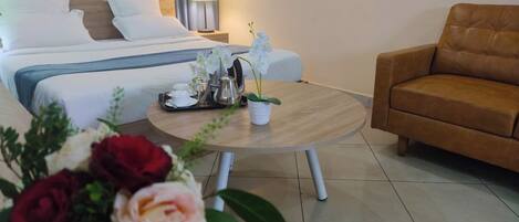 Quarto de hotel com cama, mesa redonda com um vaso de flores, sofá marrom e mesa de cabeceira com abajur.