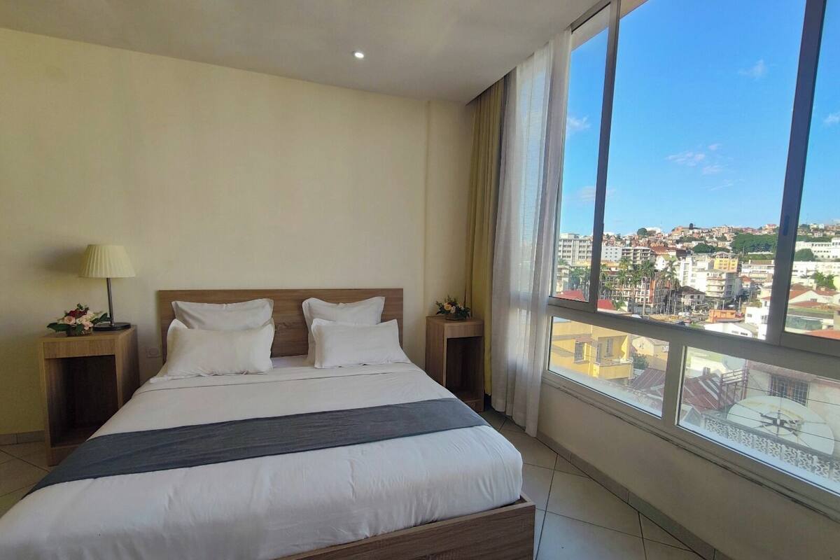 Een hotelkamer met een groot bed, twee nachtkastjes en uitzicht op de stad door grote ramen.