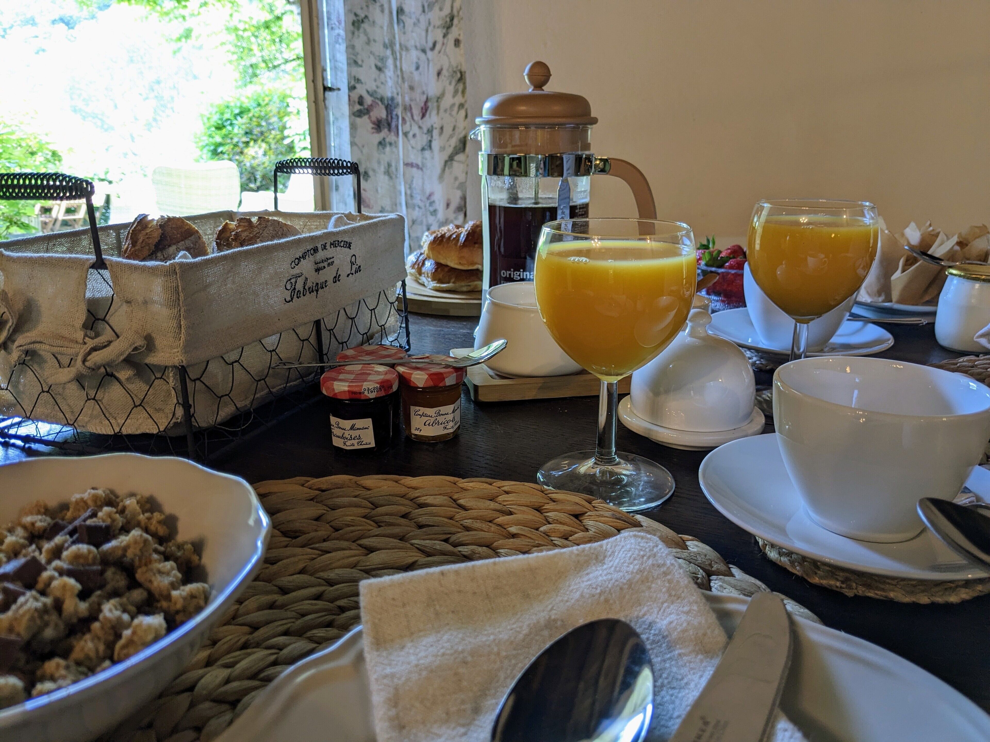 A breakfast setting with a coffee pot, two glasses of orange juice, a basket of bread, and a bowl of cereal.