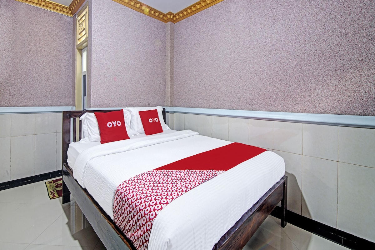 A hotel room with a bed, white bedding, red pillows, and a red and white patterned throw.