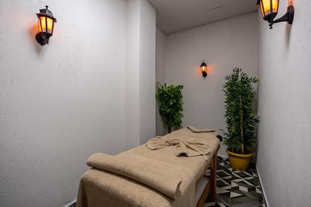 A small, narrow room with a massage table covered in a beige towel, two wall-mounted lamps, and two potted plants.