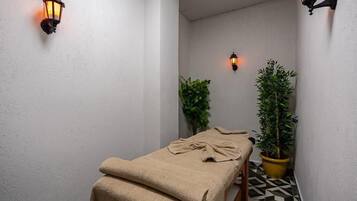 A small, narrow room with a massage table covered in a beige towel, two wall-mounted lamps, and two potted plants.