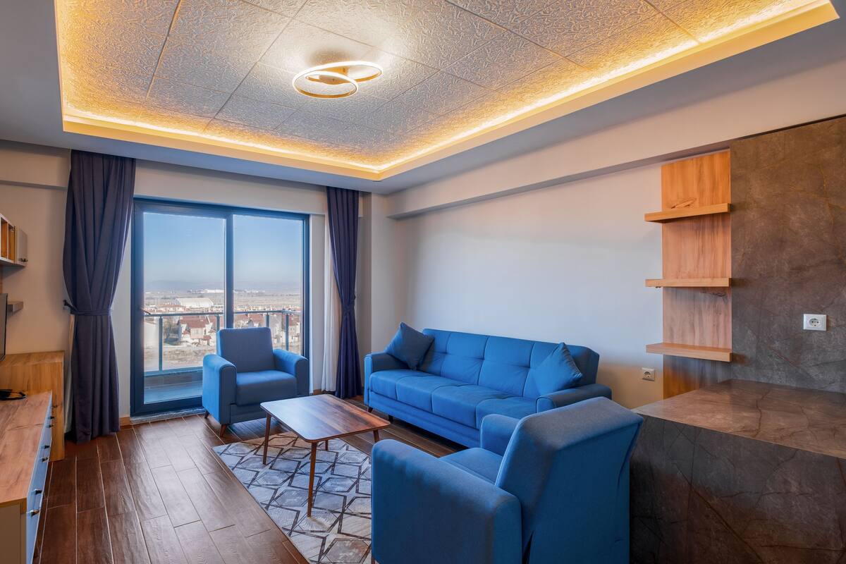 A modern living room with a blue sofa, two armchairs, a wooden coffee table, and a large window offering a city view.