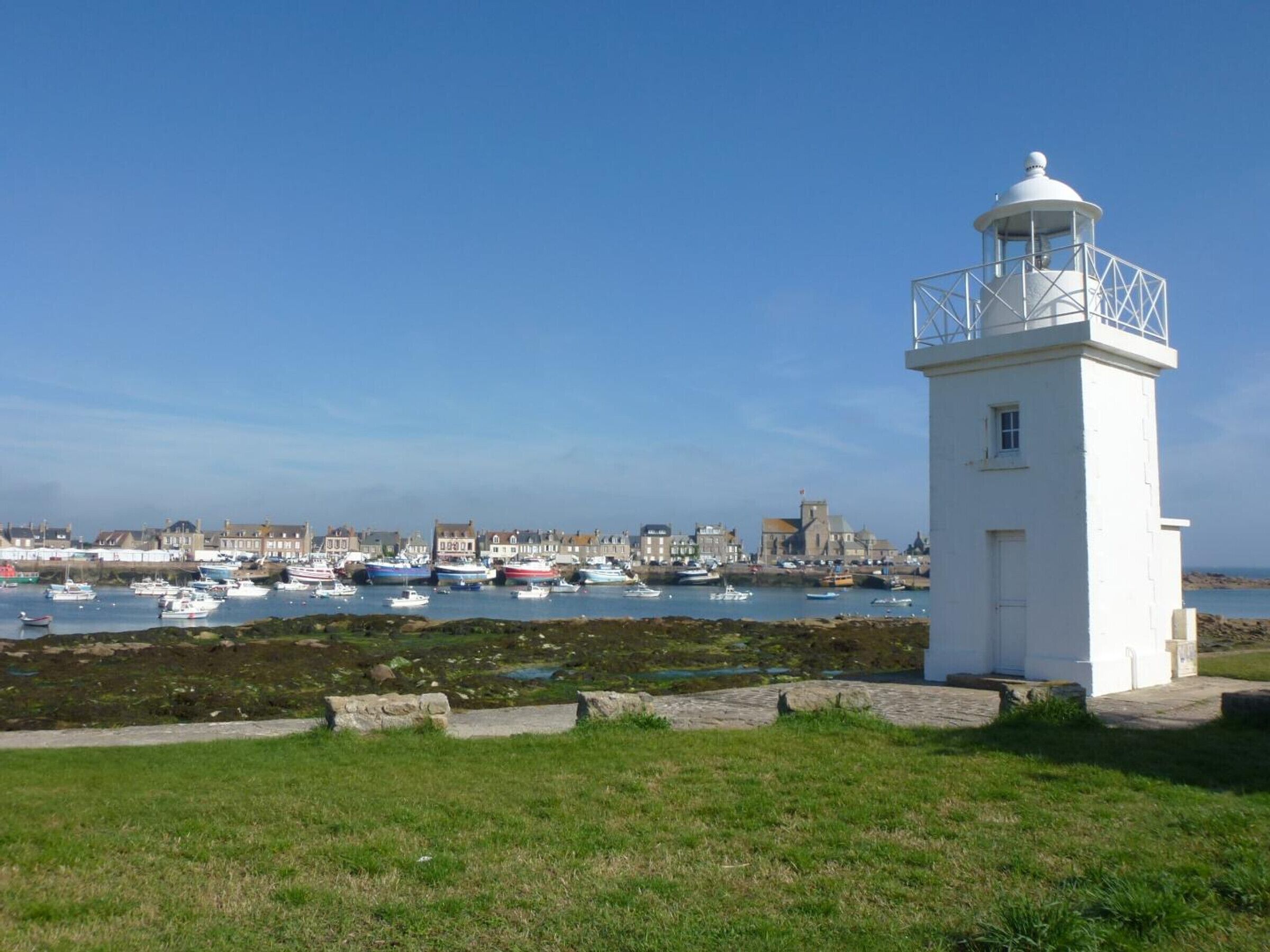 A white lighthouse with a balcony, a grassy foreground, and a marina with boats.