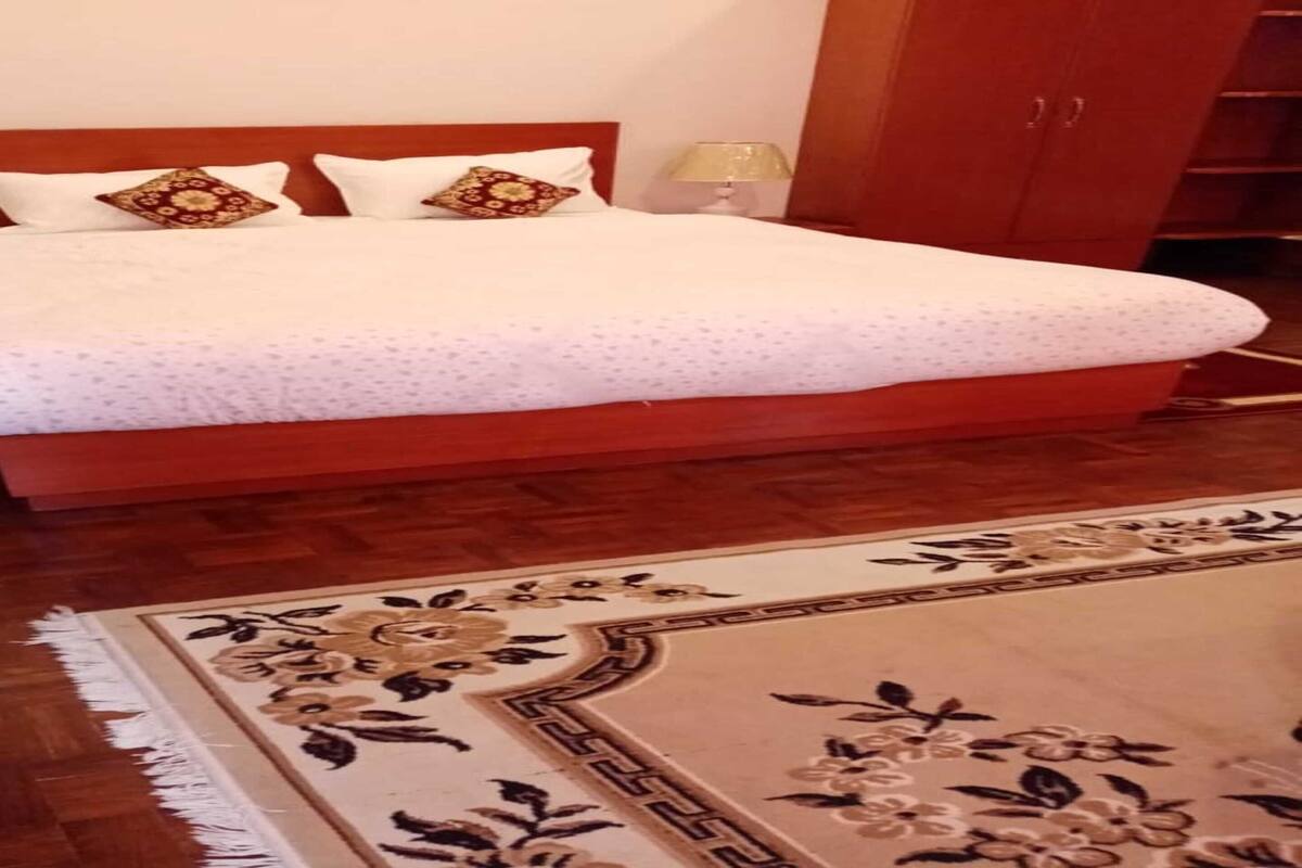A bedroom with a wooden bed, white bedding, a floral-patterned rug, and a potted plant.