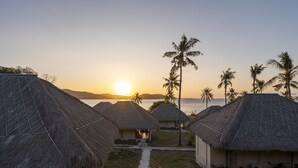 Casette dal tetto di paglia con un sentiero che conduce alla spiaggia, palme e l'oceano sullo sfondo.