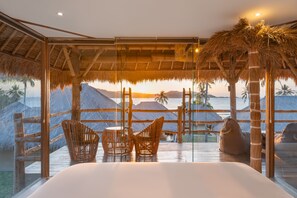 Une chambre dotée d’un grand lit, de fauteuils en osier et offrant une vue sur l’océan au coucher du soleil.