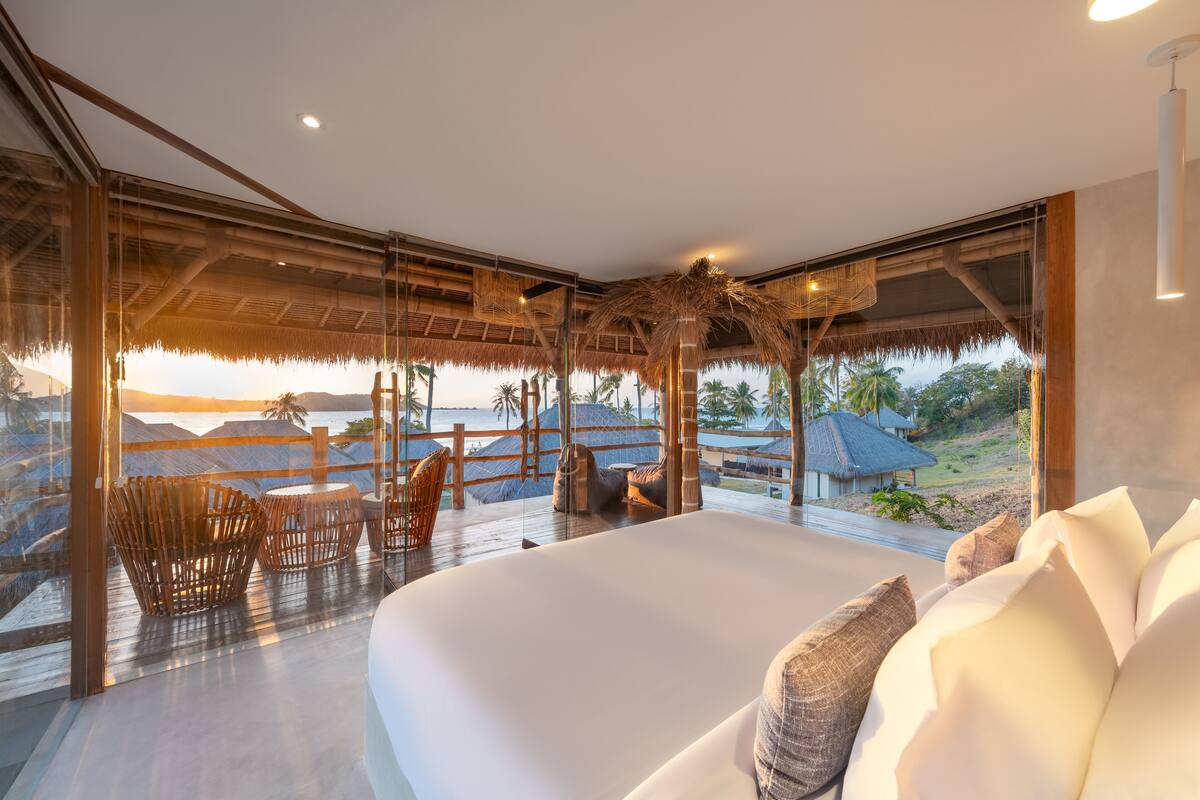 A modern bedroom with a large bed, a glass wall, and a view of the beach.