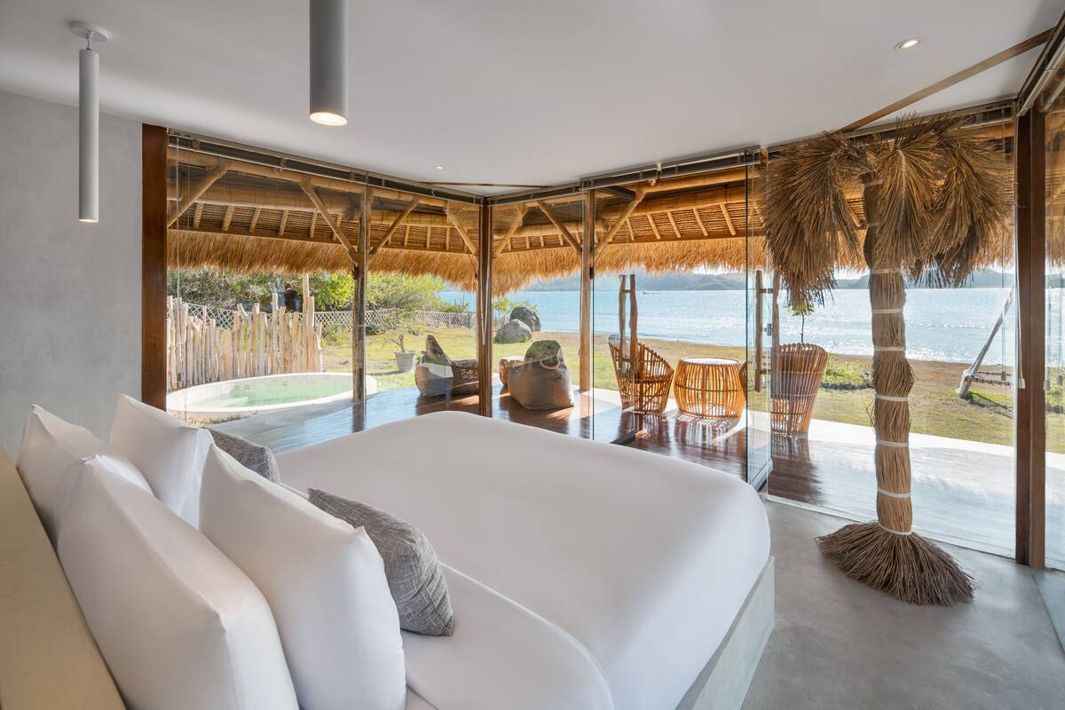 A modern bedroom with a large bed, a view of the sea, and a thatched roof outside.